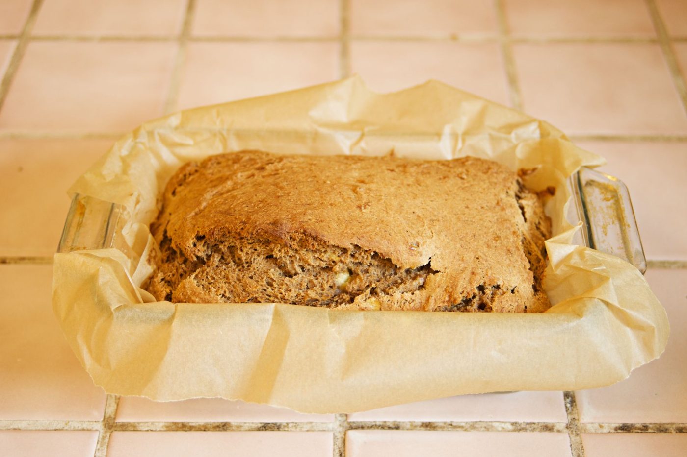 A gluten free banana nut bread loaf partially wrapped in plastic wrap on a kitchen counter, with a few slices on a plate